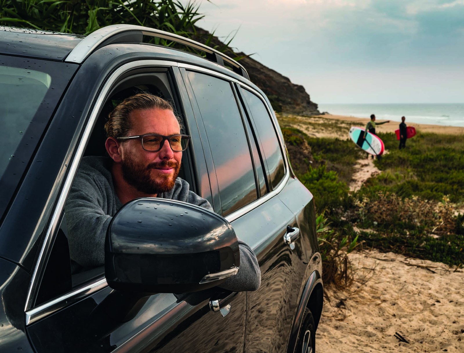 A smiling man in glasses leaning from the window of his car by the beach
